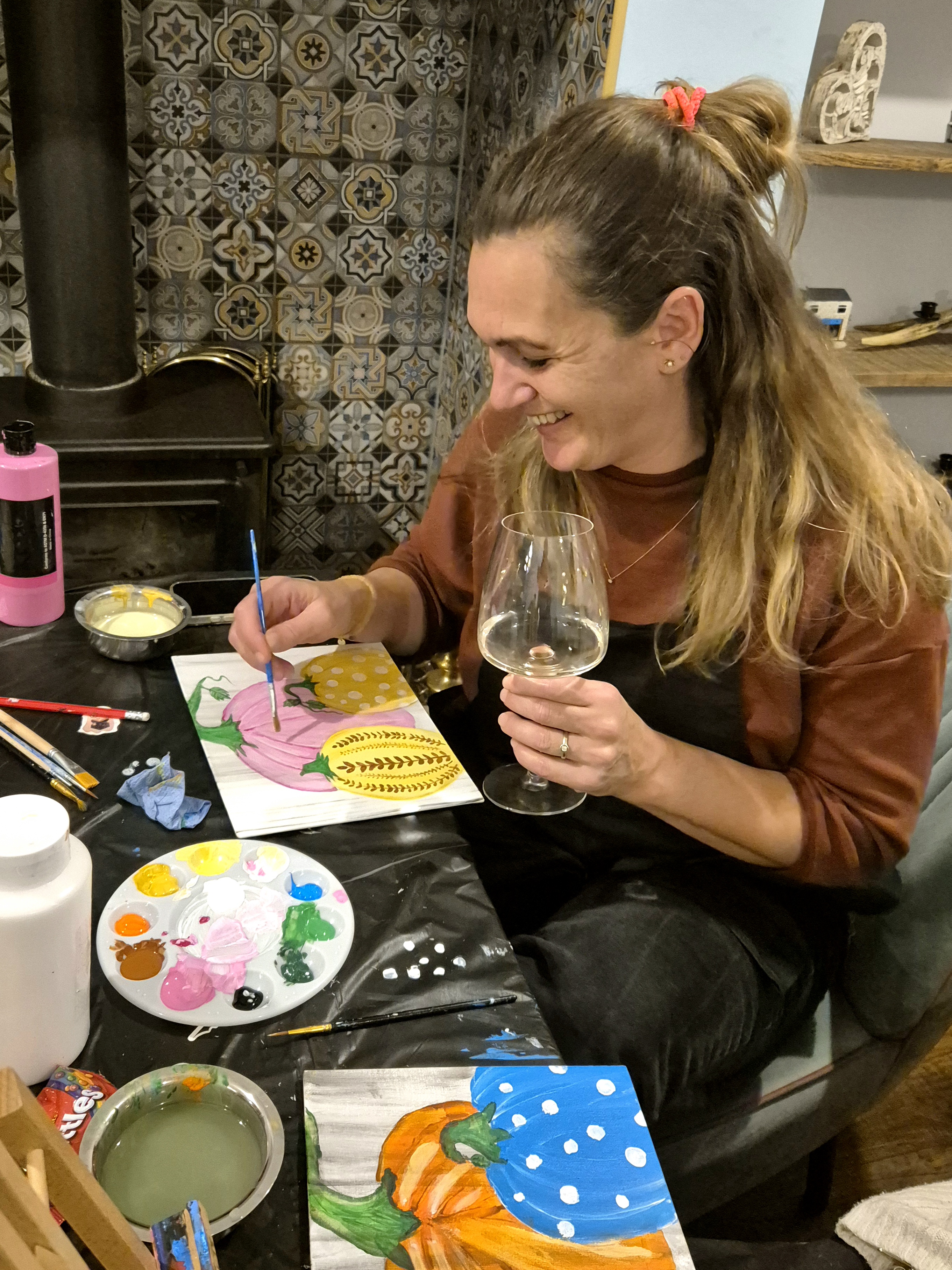Photo of a person painting while holding a glass of wine. They are smiling!