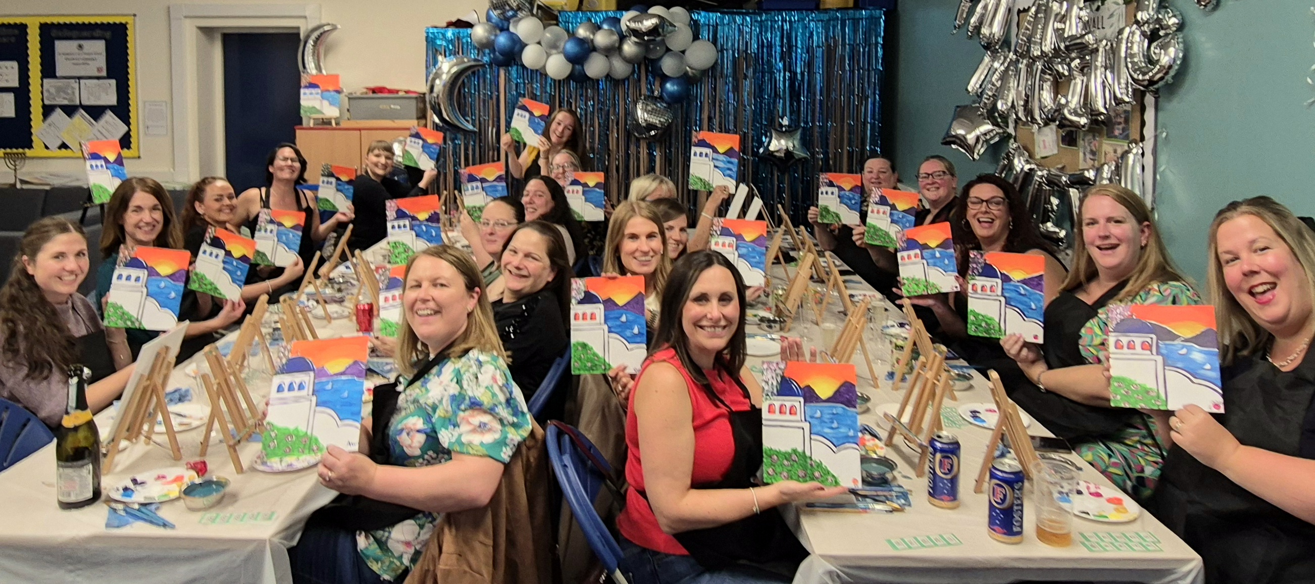 Photo of a large group of people seated at two long tables. They are all holding up paintings.