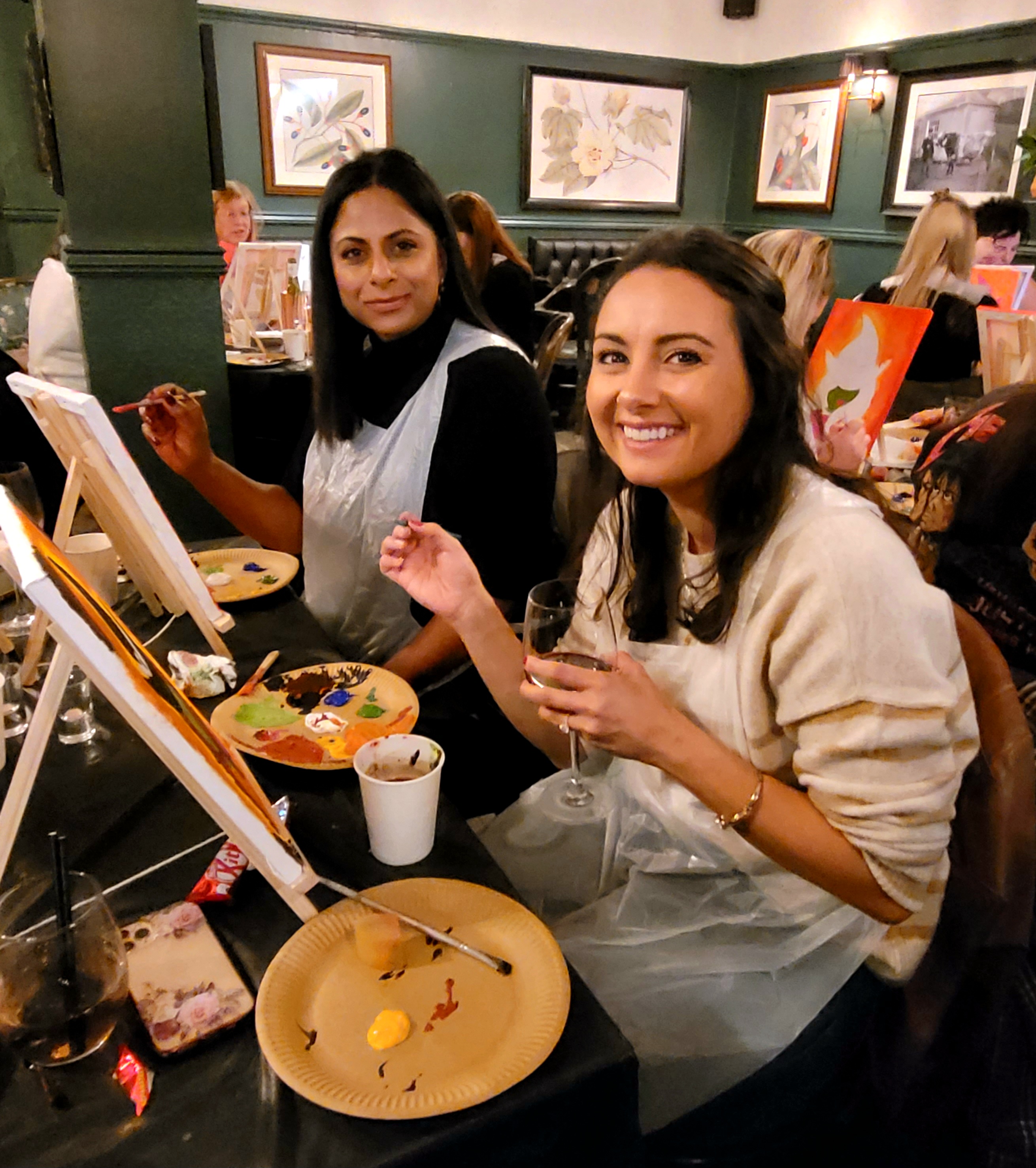 Picture of 2 people at a Paint and Sip event.  One is holding a wine glass and paintbrush, the other a paintbrush.  They are both smiling at the camera.