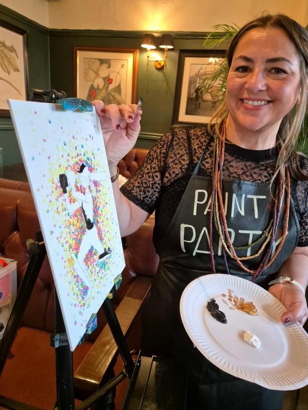 Photo of a woman stood at a paint easel holding a brush and a palette.  She is smiling at the camera.